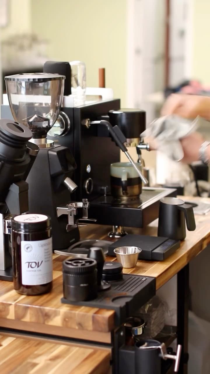 Close up of the bar in service: a black grinder and espresso machine on a butcher-block top, a jar of Tov coffee in the foreground, a milk pitcher and scale to one side.