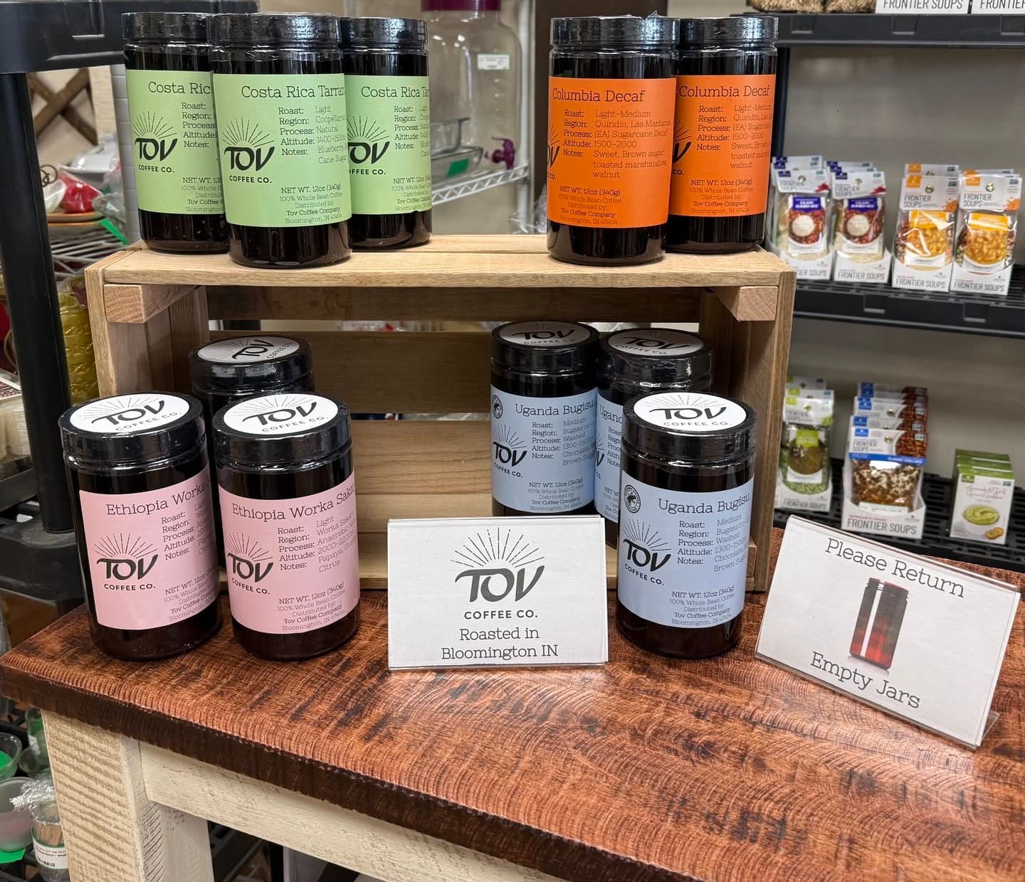 Tov jars on a wooden riser in a shop: Costa Rica Tarrazu, Colombia Decaf, Ethiopia Worka Sakaro and Uganda Bugisu, each with a colored label, beside a card reading Please Return Empty Jars.